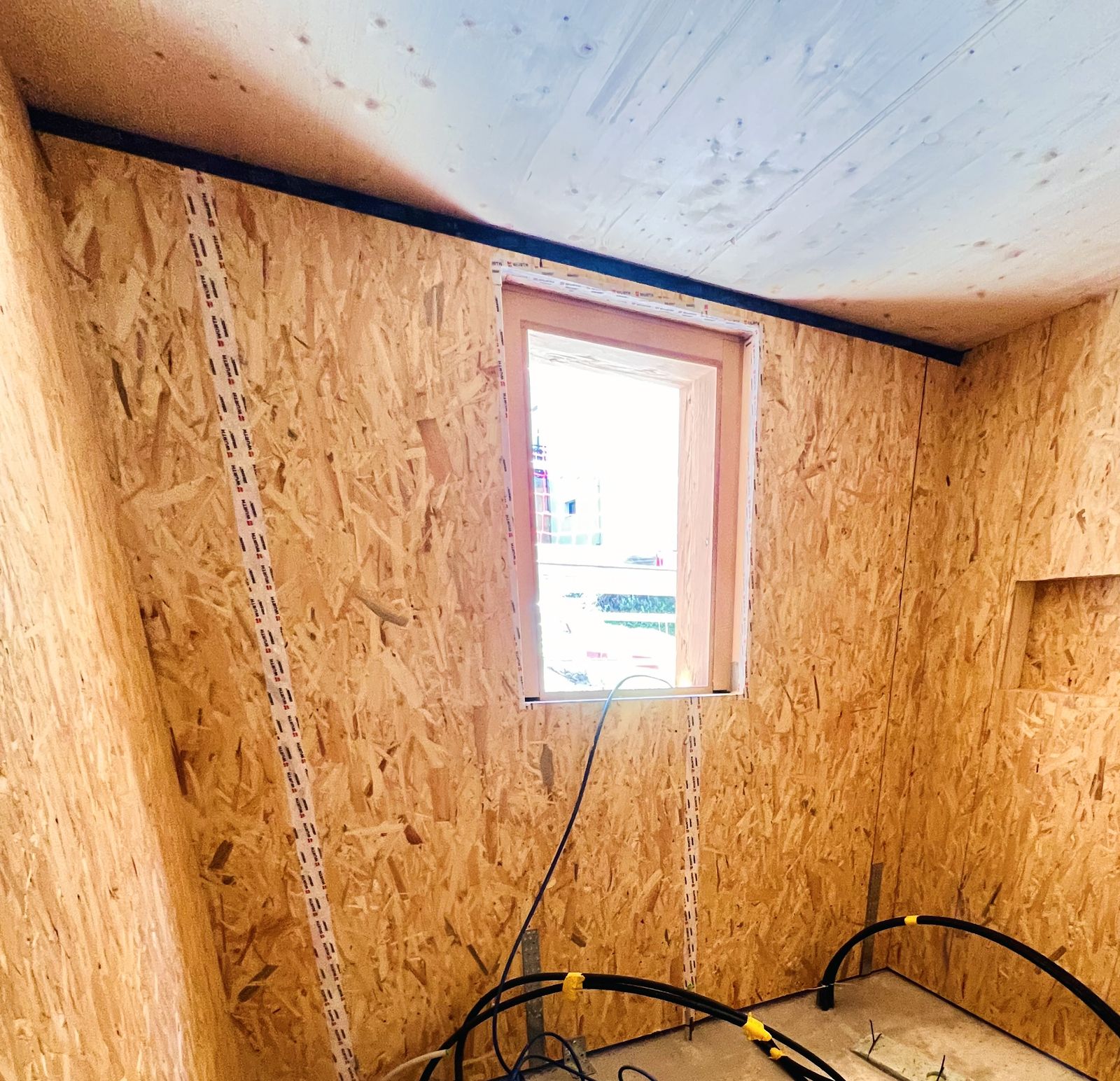 Lato interno di una parete a telaio in legno durante il montaggio, con pannello OSB, giunti nastrati e raccordo della finestra nastrato sotto un solaio in legno