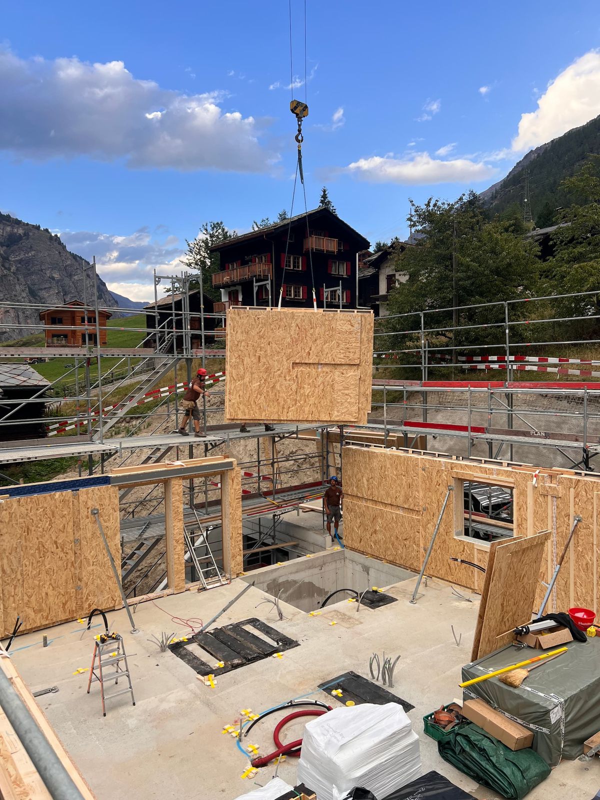 Kran hebt ein vorgefertigtes Wandelement auf einer Baustelle in Randa VS, im Hintergrund Walliser Chalets